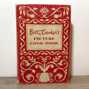 May include: Vintage cookbook titled "Betty Crocker's Picture Cook Book." The cover is red with an ornate cream-colored floral design. The title is in a cream-colored rectangular box. The book appears to be in good condition.