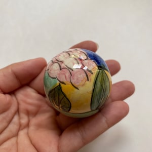 Peut inclure: Une petite boule en céramique ronde avec un motif peint à la main de fleurs roses et de feuilles vertes sur un fond jaune, bleu et vert.
