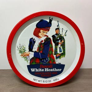 May include: Red and white tin with a red rim. The tin features a woman and a man in traditional Scottish attire. The woman is holding a sprig of heather. The text on the tin reads "Pascall White Heather Chocolates and Toffees Net Wt. 15 1/2 Oz. 439 g."