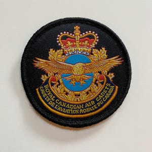 May include: A round patch with a black border displaying the Royal Canadian Air Cadets emblem. The design includes a golden eagle, a crown, and text in both English and French, set against a blue background.