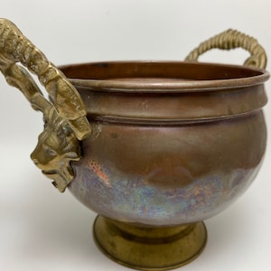 May include: A copper planter with two ornate brass handles shaped like lion heads. The planter has a round base and a slightly flared top.