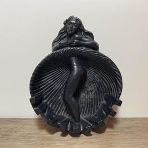 May include: A black sculpture of a woman with long hair, wearing a dress that fans out in a circular shape. The sculpture is set against a white background and rests on a wooden surface.