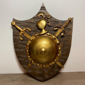 May include: A decorative wooden shield with a gold-colored central emblem featuring a knight's helmet and a circular shield. Two gold swords are crossed behind the shield. The shield has a dark brown, textured background.