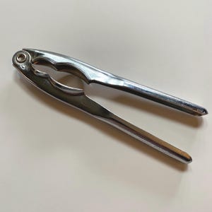 May include: A shiny, silver nutcracker with a classic design. The tool has a hinged mechanism with curved handles and a pointed tip. The nutcracker is made of metal and is designed for cracking nuts.