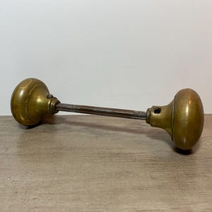 Peut inclure: Une poignée de porte ancienne en laiton, avec une longue tige filetée. Les boutons sont arrondis et présentent une teinte dorée chaude. Le métal montre des signes d'âge et d'usure, suggérant une pièce vintage.