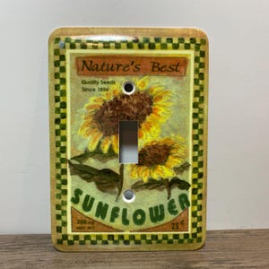 May include: A decorative light switch cover featuring a sunflower design. The cover has a rectangular shape with rounded corners and a checkered border. The words "Nature's Best" and "Sunflower" are printed on the cover.