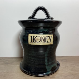 May include: A dark blue ceramic honey pot with a lid and handle. The pot features a cream-coloured label with the word "HONEY". The jar has a distinctive shape and a glossy finish. The jar is suitable for storing honey.