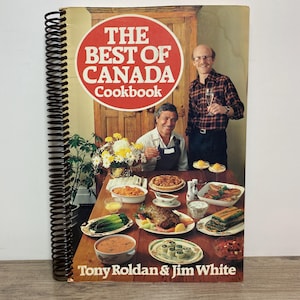 May include: A cookbook titled "The Best of Canada Cookbook" with a red and white circular logo. The cover features two men, a table laden with food, and the names Tony Roldan & Jim White. The book has a spiral binding.