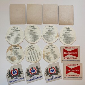 May include: A collection of vintage beer coasters and promotional items. The coasters feature various designs, including the Budweiser logo, the text "Make it a Kind of Day", and the Moosehead Brewery logo. Some coasters are round, others square.