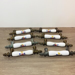 May include: A set of eight drawer pulls featuring white ceramic handles with a colorful floral pattern. Each handle is flanked by aged brass metal hardware with a decorative, tiered finial design on both ends, showing signs of oxidation.
