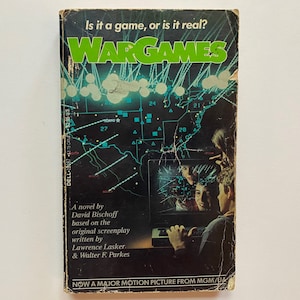 May include: A book cover with the title "WARGAMES" in green letters. The cover features a map of the United States with red and blue dots and lines. The text "Is it a game, or is it real?" is at the top of the cover. The text "A novel by David Bischoff based on the original screenplay written by Lawrence Lasker & Walter F. Parkes" is at the bottom of the cover. The text "NOW A MAJOR MOTION PICTURE FROM MGM/UA" is at the bottom of the cover.