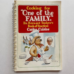 May include: A red and white cookbook with a spiral binding. The cover features a cartoon dog wearing a chef's hat and apron, holding a can of dog food. The title of the book is "Cooking for "One of the FAMILY." Dr. Bernard Tonken's Book of Practical Canine Cuisine."