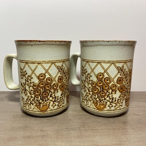 以下が含まれることがあります： 白い背景に茶色の花柄が描かれたセラミック製のマグカップ2個。マグカップは茶色の縁取りと持ち手があります。