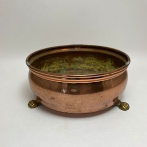 May include: A copper planter with a rounded shape and three brass feet. The planter has a slightly aged patina.