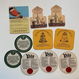 May include: A collection of vintage beer coasters and promotional cards. The coasters feature various designs, including a sailboat, cocktails, and bell illustrations. Some coasters have text, including "Captain Morgan Caribbean Competition" and "Pils From Holsten."
