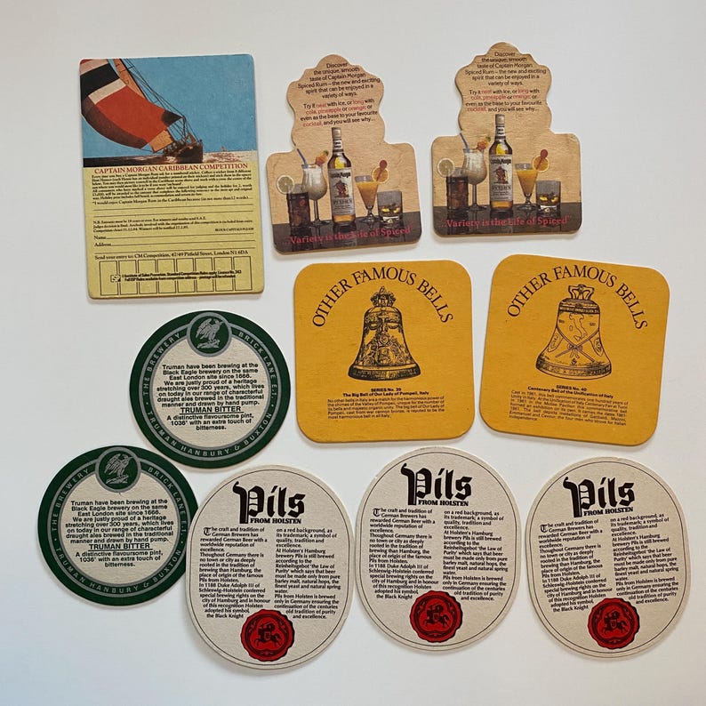 May include: A collection of vintage beer coasters and promotional cards. The coasters feature various designs, including a sailboat, cocktails, and bell illustrations. Some coasters have text, including "Captain Morgan Caribbean Competition" and "Pils From Holsten."