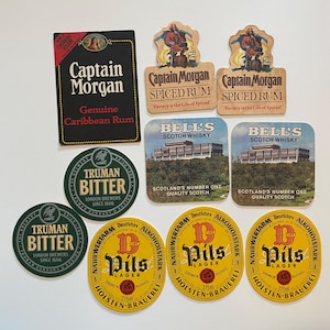 May include: A collection of vintage drink coasters featuring various brands. Includes Captain Morgan Spiced Rum, Bell's Scotch Whisky, Truman Bitter, and Holsten-Brauerei Pils Lager. The coasters are in various shapes and colors.
