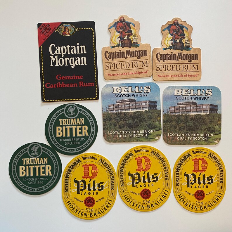 May include: A collection of vintage drink coasters featuring various brands. Includes Captain Morgan Spiced Rum, Bell's Scotch Whisky, Truman Bitter, and Holsten-Brauerei Pils Lager. The coasters are in various shapes and colors.