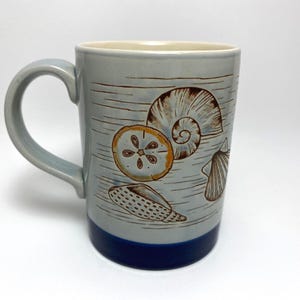 May include: A ceramic mug with a light blue glaze and a dark blue base. The mug features illustrations of seashells in shades of brown and orange. The handle is light blue, and the interior is a cream colour. The mug is designed for hot beverages.