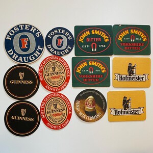 May include: A collection of vintage beer coasters featuring logos for Foster's Draught, Guinness, John Smith's Bitter, Hofmeister, and Henry Wintermans. The coasters are round and rectangular, with various colors and designs.