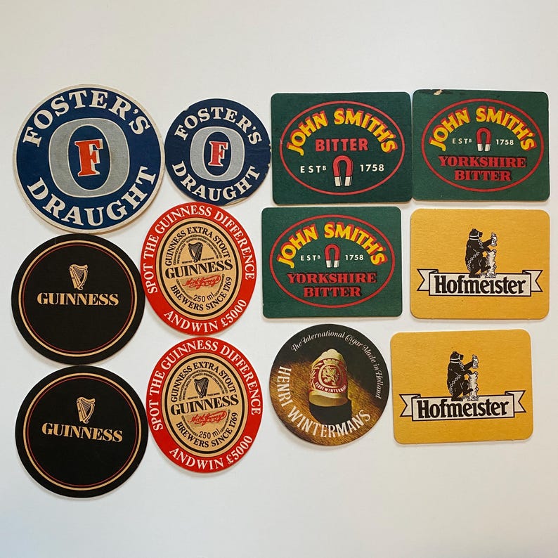 May include: A collection of vintage beer coasters featuring logos for Foster's Draught, Guinness, John Smith's Bitter, Hofmeister, and Henry Wintermans. The coasters are round and rectangular, with various colors and designs.