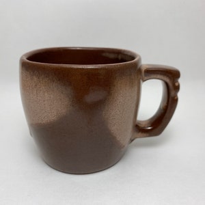 以下が含まれることがあります： 正方形のトップと丸みを帯びたボディを備えた茶色のセラミックマグ。マグカップにはハンドルがあり、2色の配色で、上半分は明るい茶色、下半分は濃い茶色です。