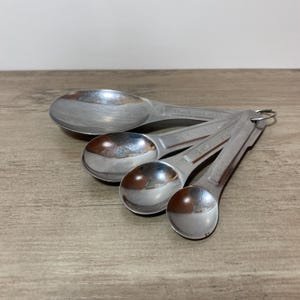 Peut inclure: Un ensemble de quatre cuillères à mesurer en acier inoxydable de différentes tailles, reliées par un anneau métallique. Les cuillères sont disposées sur une surface en bois, la plus grande étant au premier plan. Des mesures sont gravées sur les manches.