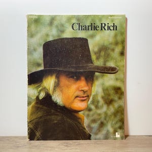 May include: A vintage album cover featuring Charlie Rich. The cover shows a portrait of a man wearing a dark hat and jacket, with the name "Charlie Rich" printed in large letters. The background is a blurred, natural scene.