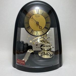 May include: A black and gold clock with a pendulum and a gold face. The clock has a clear glass dome and the words "Rhythm Quartz" on the face.