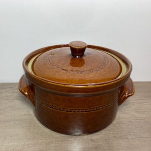 May include: A brown ceramic casserole dish with a lid. The dish has two handles and a decorative band around the middle. The lid has a knob and a decorative pattern. The interior rim is a lighter colour.