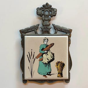 Peut inclure: Ancien dessous de plat en métal avec une incrustation de carreau de céramique. Le carreau représente une femme en robe turquoise portant une baguette, avec des épis de blé et une gerbe de blé. Le cadre en métal a un design orné.