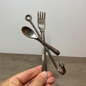 May include: A silver-colored metal wall hook featuring a fork, spoon, and a small circular element. The fork and spoon are crossed, with the hook curving downwards. The design is a unique kitchen or dining room decor item.