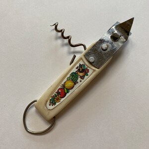 May include: A white multi-tool with a corkscrew, bottle opener, and a ring on the end. The handle is decorated with a colourful design of vegetables and flowers.