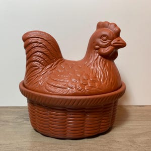 May include: A terracotta-coloured chicken-shaped container with a basket-weave base. The lid is a detailed sculpture of a chicken. The container is likely used for storing eggs or as a decorative kitchen item. The item is made of ceramic.