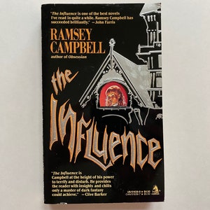May include: A book cover with a black background and a silver and orange title "The Influence" by Ramsey Campbell, author of Obsession. The cover features a silver illustration of a house with a window that shows a girl looking out.