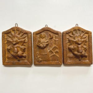 May include: Three brown wooden wall plaques with relief carvings. The plaques feature different scenes: a sun with a cherub, a woman with a building, and another sun with a cherub. Each plaque has a metal loop for hanging.