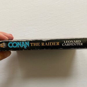 Może przedstawiać: Czarna książka w miękkiej oprawie z tytułem "Conan the Raider" autorstwa Leonarda Carpentera. Książka została wydana przez Tor Books.