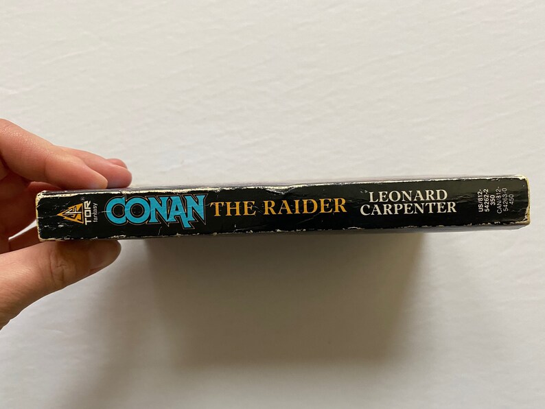 Może przedstawiać: Czarna książka w miękkiej oprawie z tytułem "Conan the Raider" autorstwa Leonarda Carpentera. Książka została wydana przez Tor Books.