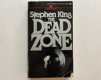 May include: A black paperback book with a silver title "The Dead Zone" by Stephen King, author of "It". The cover features a black and white image of a man's face with a clock face behind it.