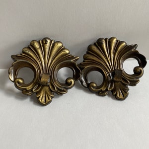 May include: Two ornate gold-toned metal hooks with a decorative floral design. The hooks are attached to a wall or surface.