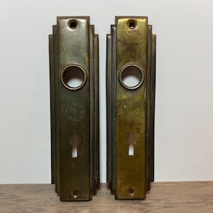 May include: A pair of antique brass door plates, each featuring a keyhole and a circular opening. The rectangular plates have Art Deco-style stepped edges. The brass has an aged, weathered patina, showing signs of use and wear.