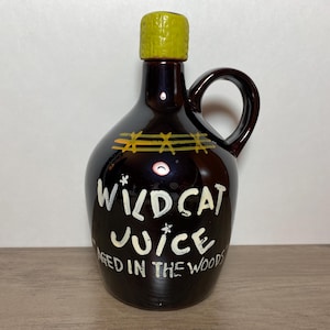 以下が含まれることがあります： 黄色の蓋と取っ手のある茶色のガラス製の壺。壺には「Wildcat Juice Aged in the Woods」という白い文字が書かれている。