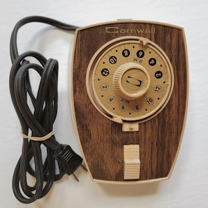 May include: A vintage Cornwall timer with a wood-grain effect. The timer has a circular dial with numbers and a central knob. It features an on/off switch and a black power cord. The timer is designed for electrical appliances.