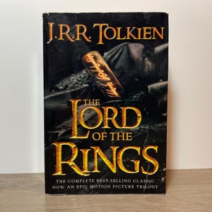 May include: A copy of "The Lord of the Rings" by J.R.R. Tolkien. The book's cover is black with gold lettering, showcasing a ring illustration. The text on the cover reads "The Complete Best-Selling Classic Now An Epic Motion Picture Trilogy."