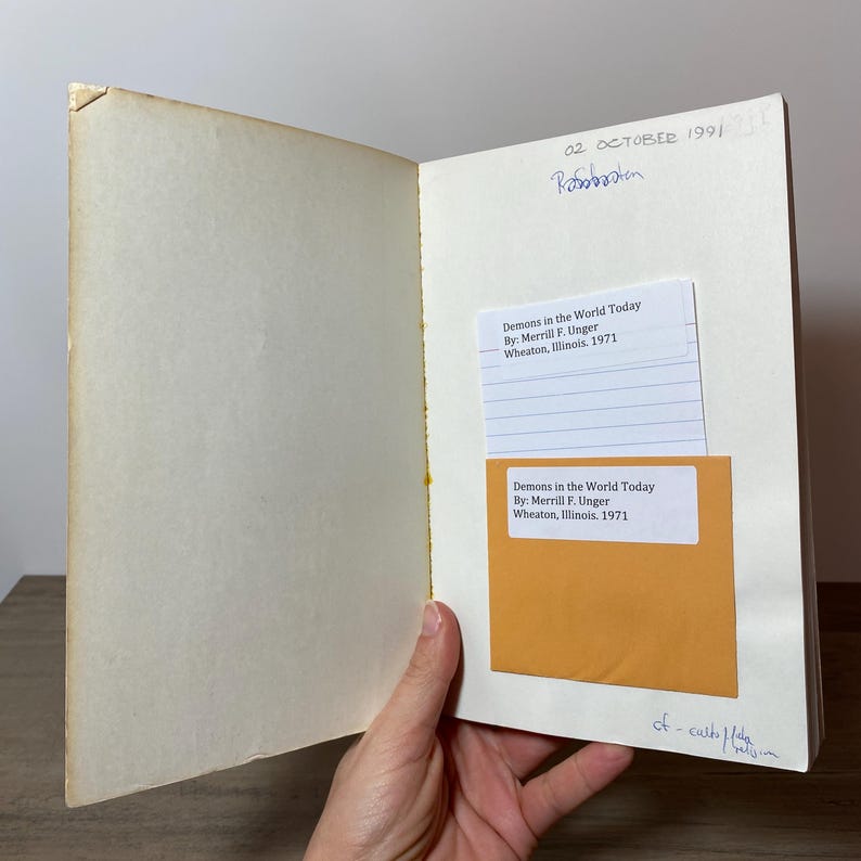 May include: An open book with aged, yellowed pages. Two labels are visible, one white and one orange, each with the text "Demons in the World Today" and author information. Handwritten text and the date "02 OCTOBER 1991" are also present.