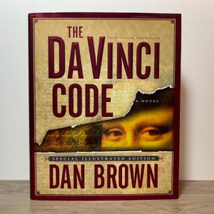 Peut inclure: Le Da Vinci Code de Dan Brown, une édition spéciale illustrée à couverture rigide. La couverture présente un fond couleur parchemin avec du texte rouge foncé, un effet de papier déchiré révélant un gros plan des yeux de la Joconde et une bordure.