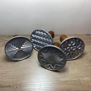 May include: Four silver metal cookie stamps with various embossed designs, including geometric patterns and starbursts. Each stamp has a small, round wooden handle. The stamps are arranged on a light brown wooden surface.