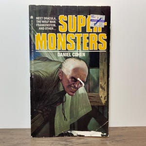 以下が含まれることがあります： ダニエル・コーエン著の「Super Monsters」というタイトルのヴィンテージペーパーバック。表紙には、黄色と白で「Meet Dracula, The Wolf Man, Frankenstein, and Other...」というテキストとともに、吸血鬼の不気味なイラストが描かれています。