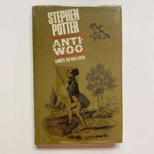 May include: A vintage book titled "Anti-Woo: Gambits for Non-Lovers" by Stephen Potter. The cover is olive green with illustrations of a man aiming a pole and a cherub with a bow. The title is in white and red.