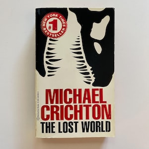 May include: A book cover featuring a black and white illustration of a dinosaur's head. The title is "The Lost World" by Michael Crichton. The cover also includes a red sticker that reads "New York Times Bestseller #1".